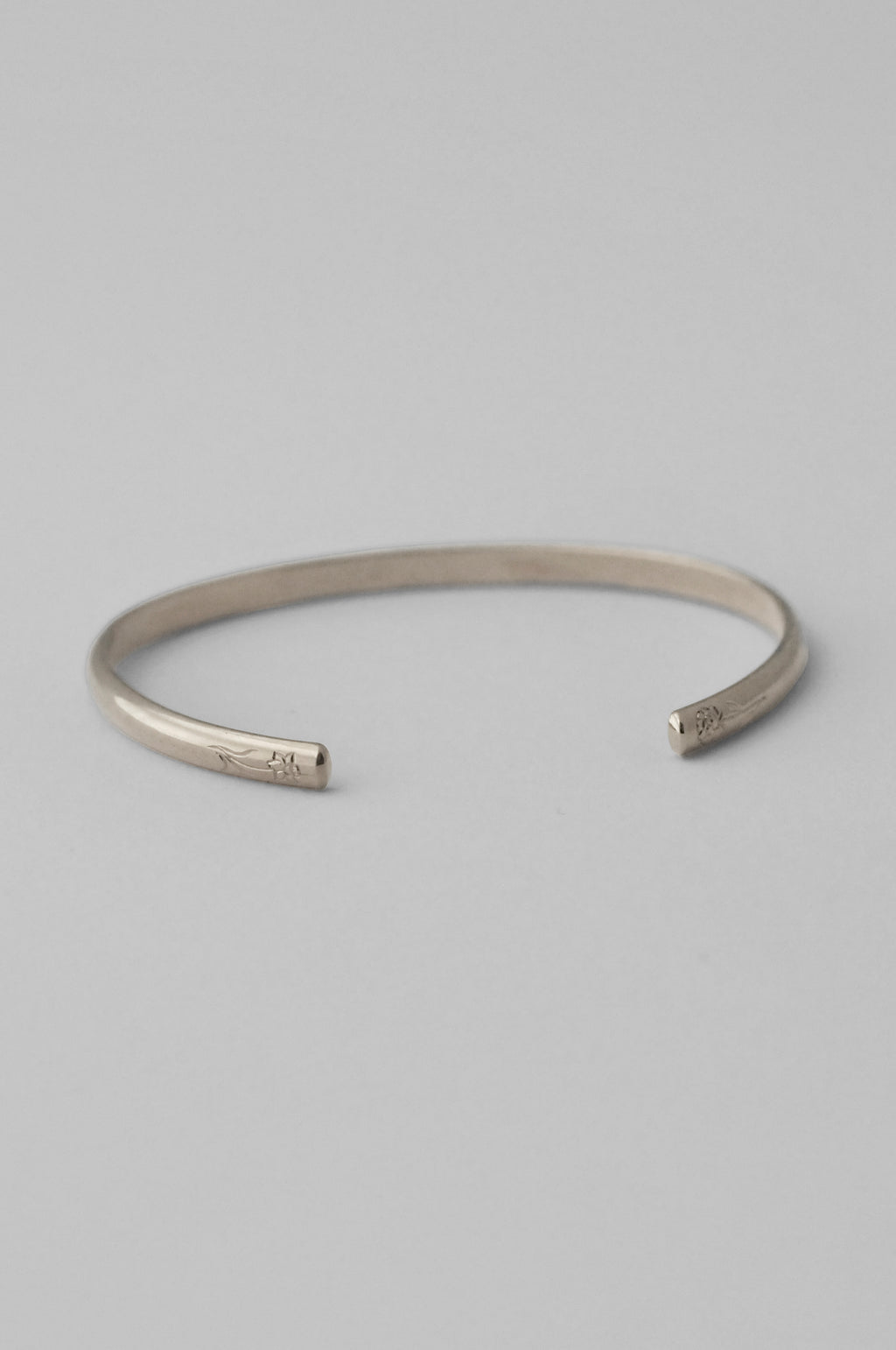 A minimalist sterling silver open cuff bangle bracelet with hand engraved flowers on each end, photographed on a clean, white background.