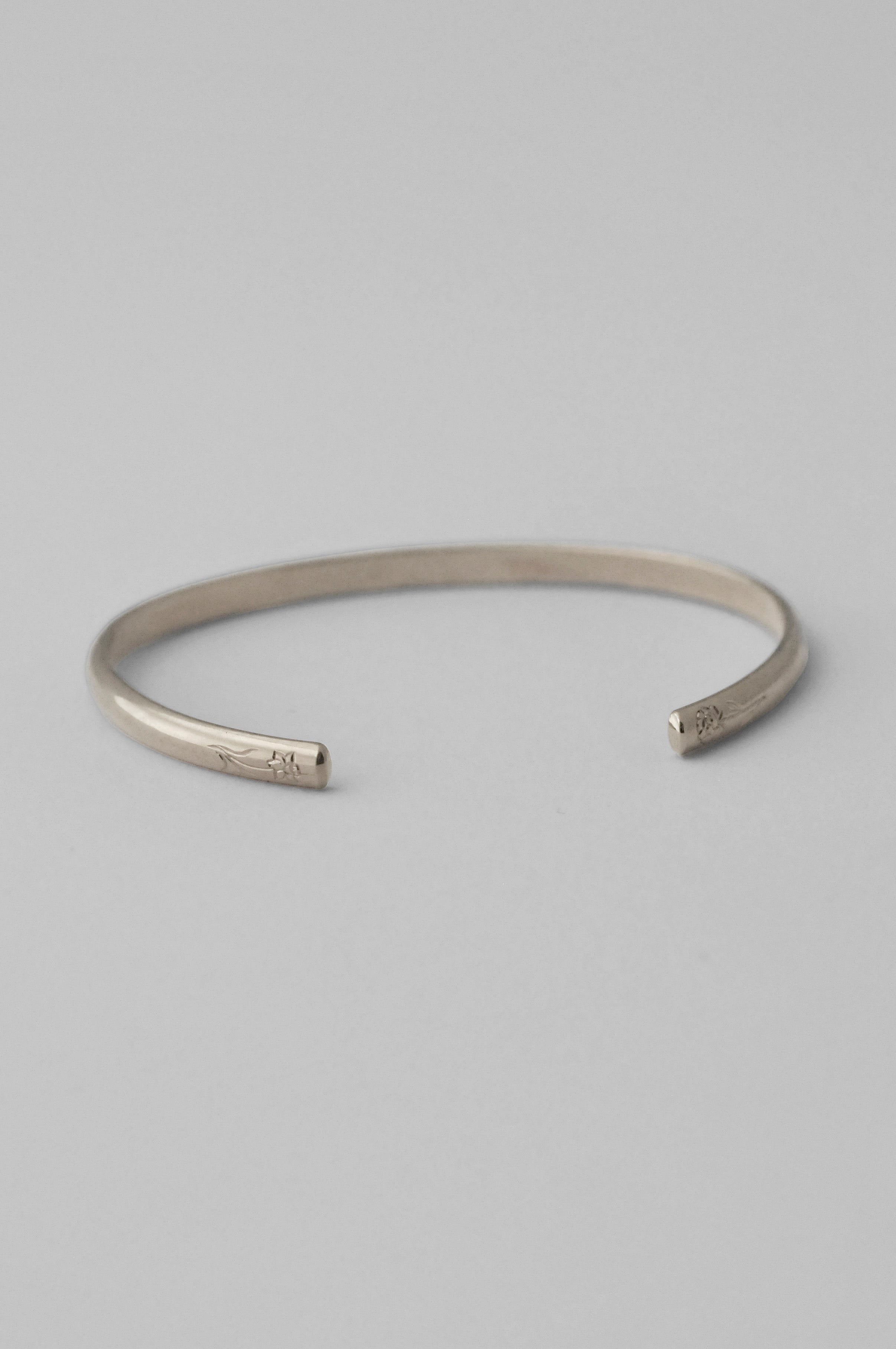 A minimalist sterling silver open cuff bangle bracelet with hand engraved flowers on each end, photographed on a clean, white background.
