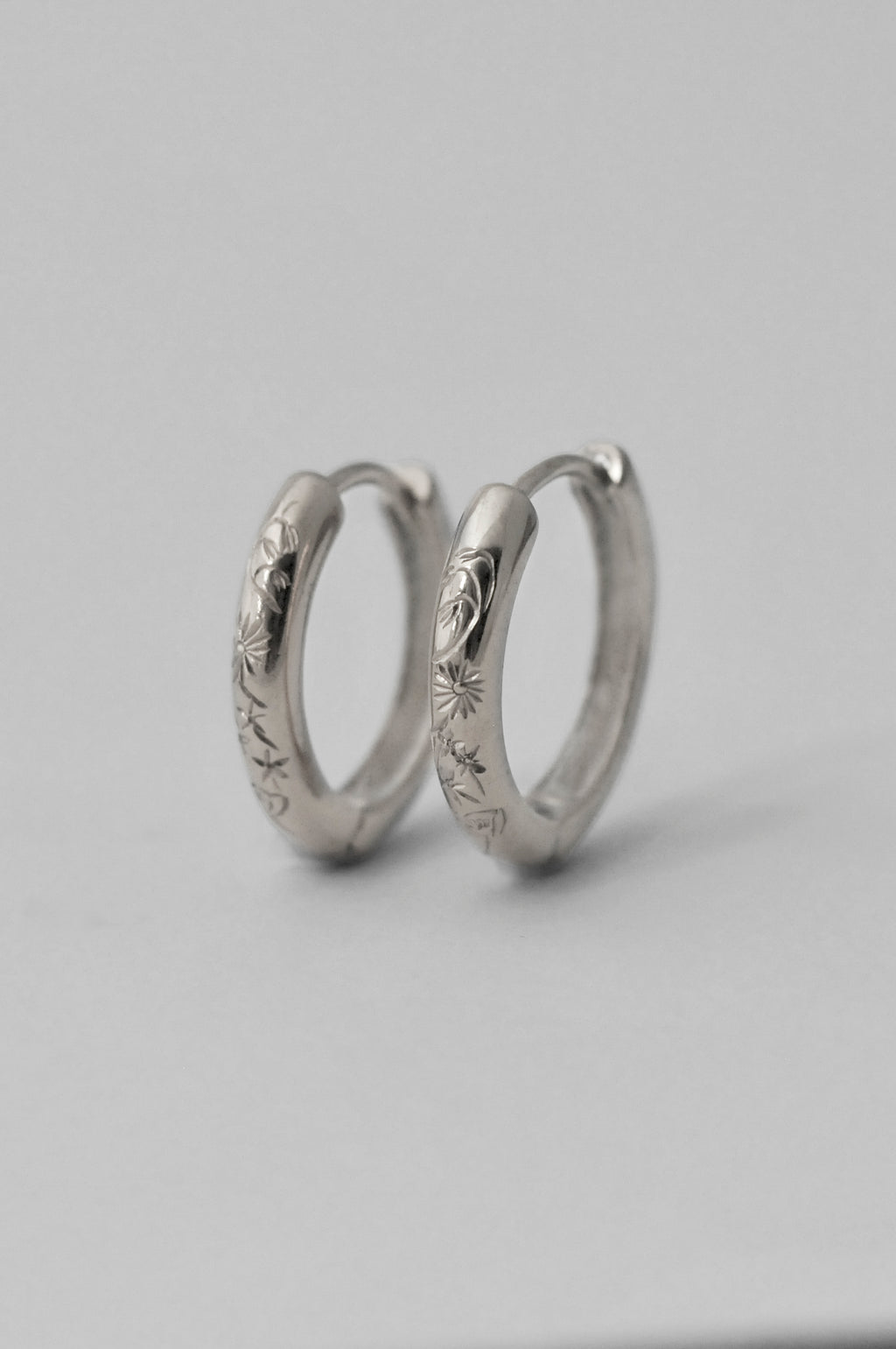 A pair of small silver hoop earrings with engraved floral patterns, displayed on a neutral light gray background.