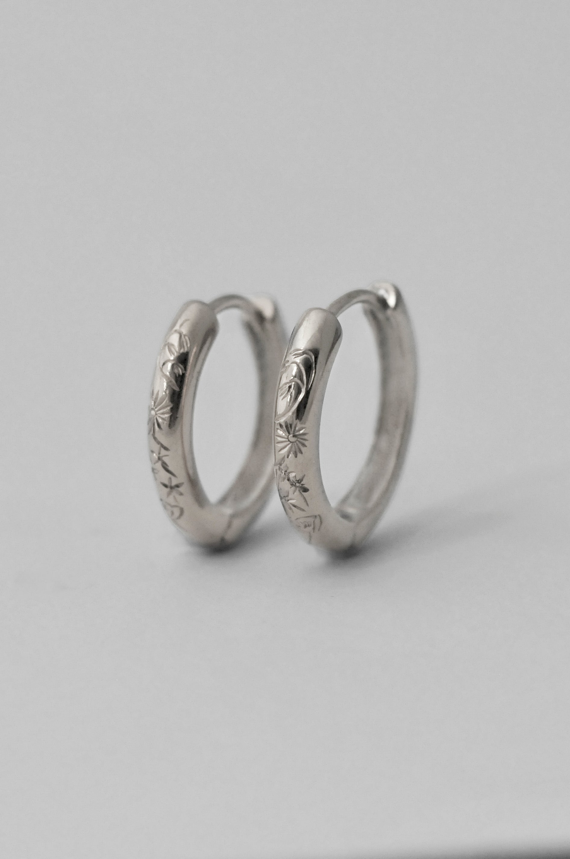 A pair of small silver hoop earrings with engraved floral patterns, displayed on a neutral light gray background.