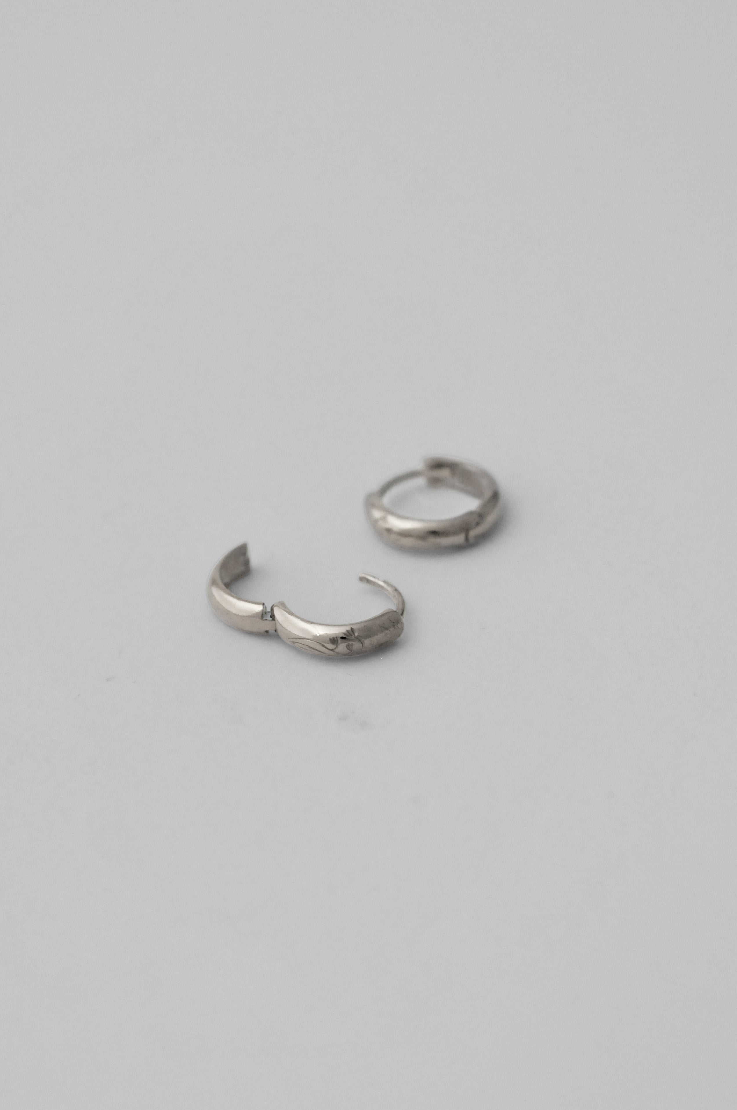 A pair of small silver huggie hoop earrings with hand engraved floral details, laying flat showing the opening of the earring, shown on a neutral gray background. 