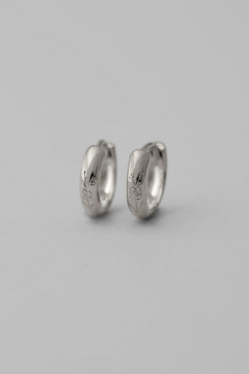 A pair of small silver huggie hoop earrings with hand engraved floral details, shown on a neutral gray background.