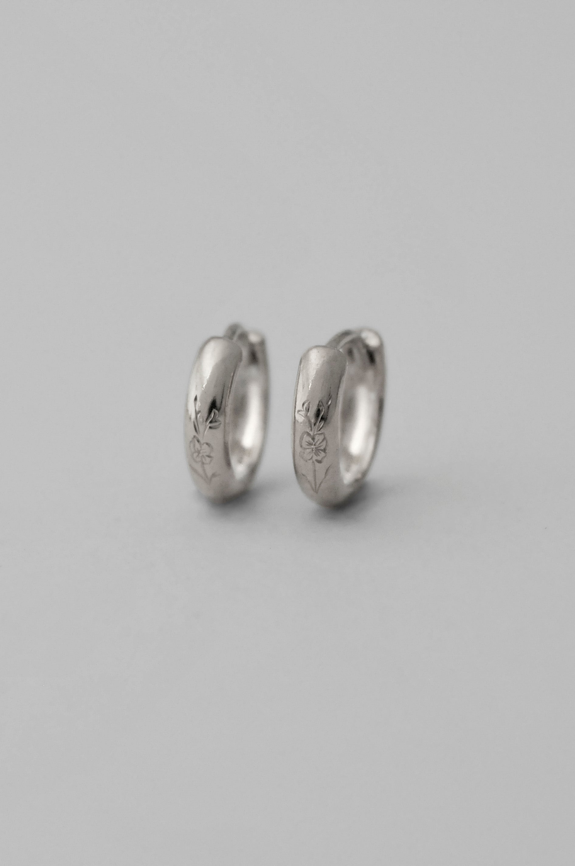 A pair of small silver huggie hoop earrings with hand engraved floral details, shown on a neutral gray background.