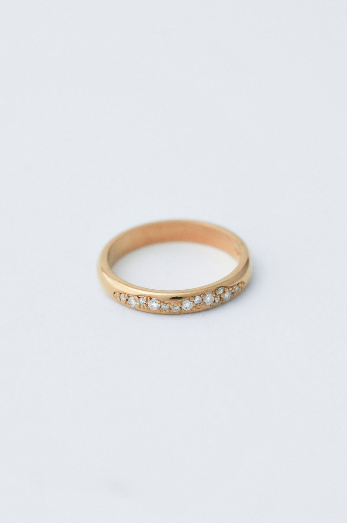 A delicate yellow gold band ring featuring a row of small clear pavé-set diamond along the front, photographed on a clean, light gray background.