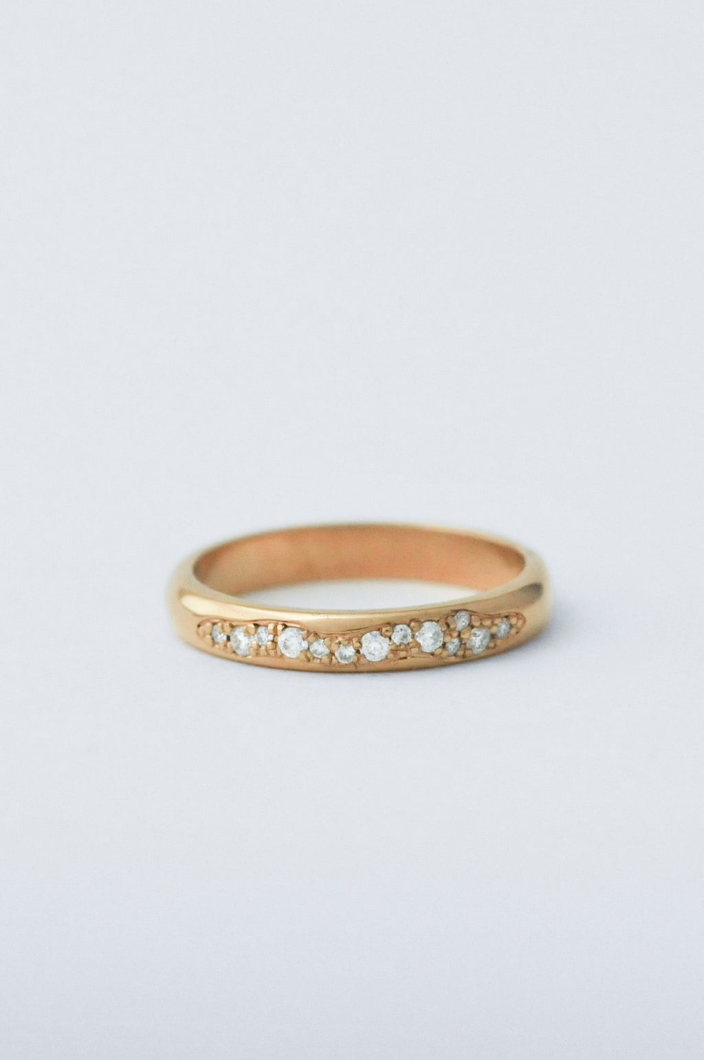 A gold band ring featuring a row of small clear round-cut diamond along the front, photographed on a clean light gray background.