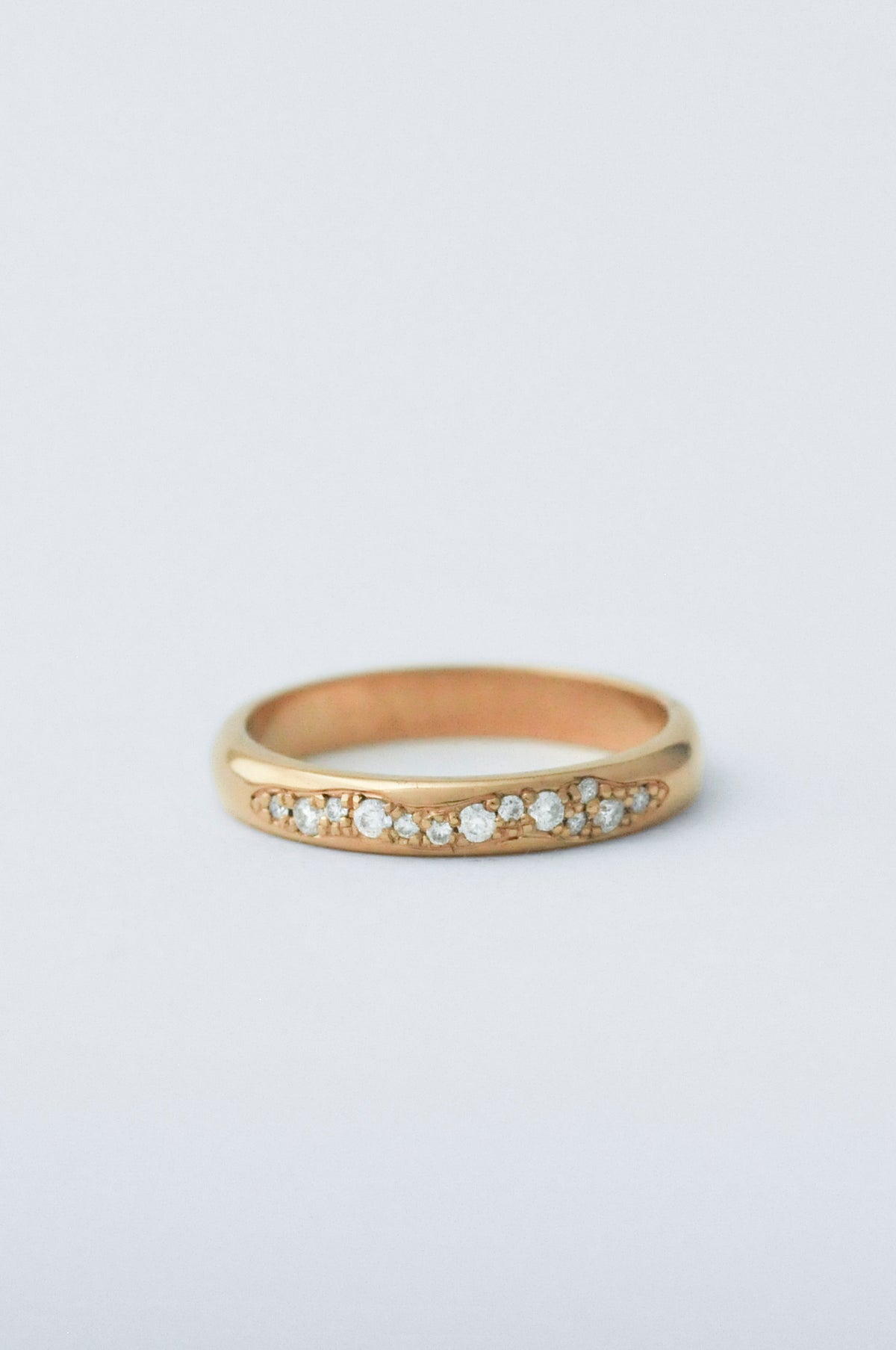 A gold band ring featuring a row of small clear round-cut diamond along the front, photographed on a clean light gray background.