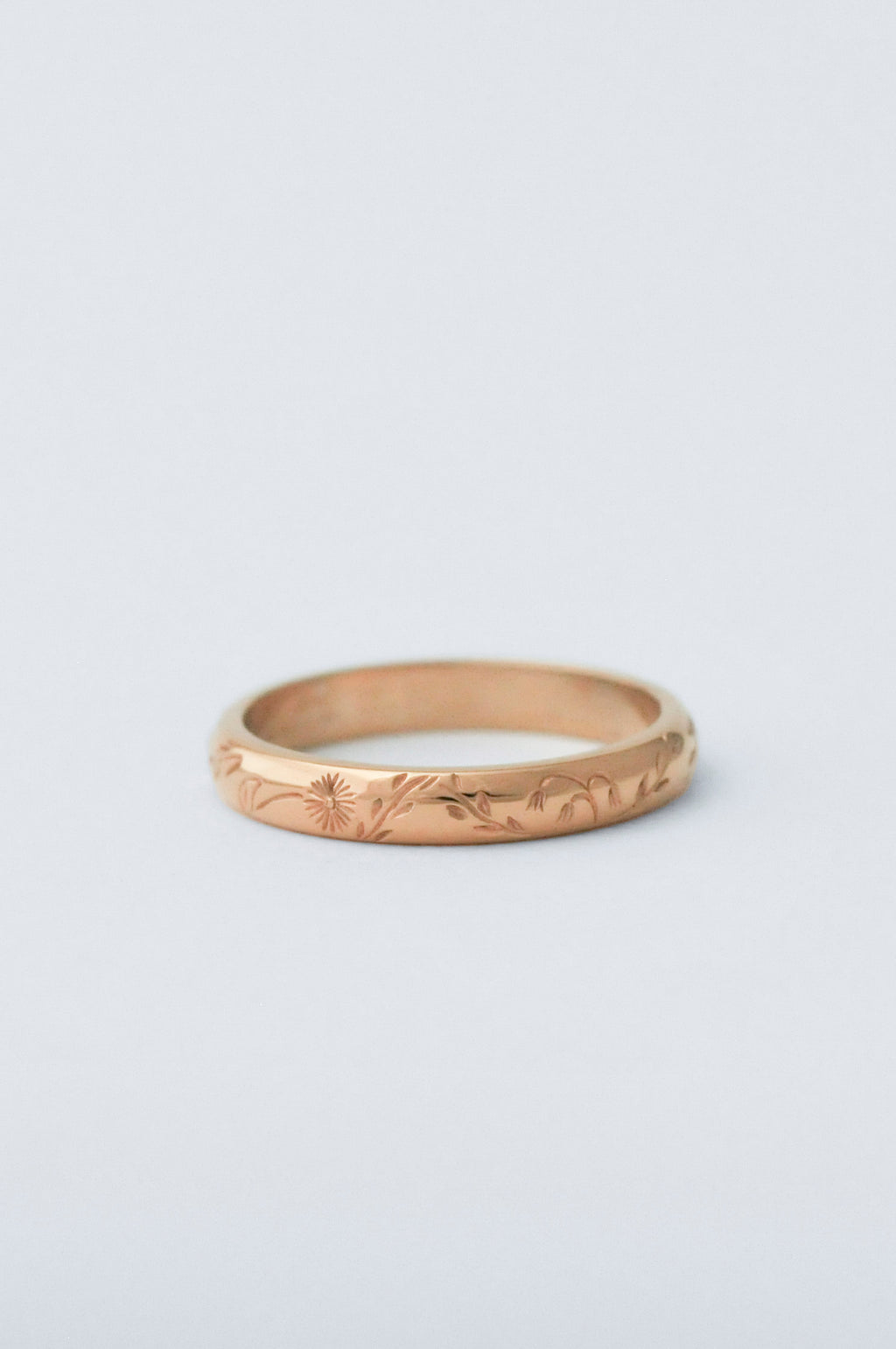 A thin gold band ring with delicate hand engraved florals, photographed on a clean, light gray background.