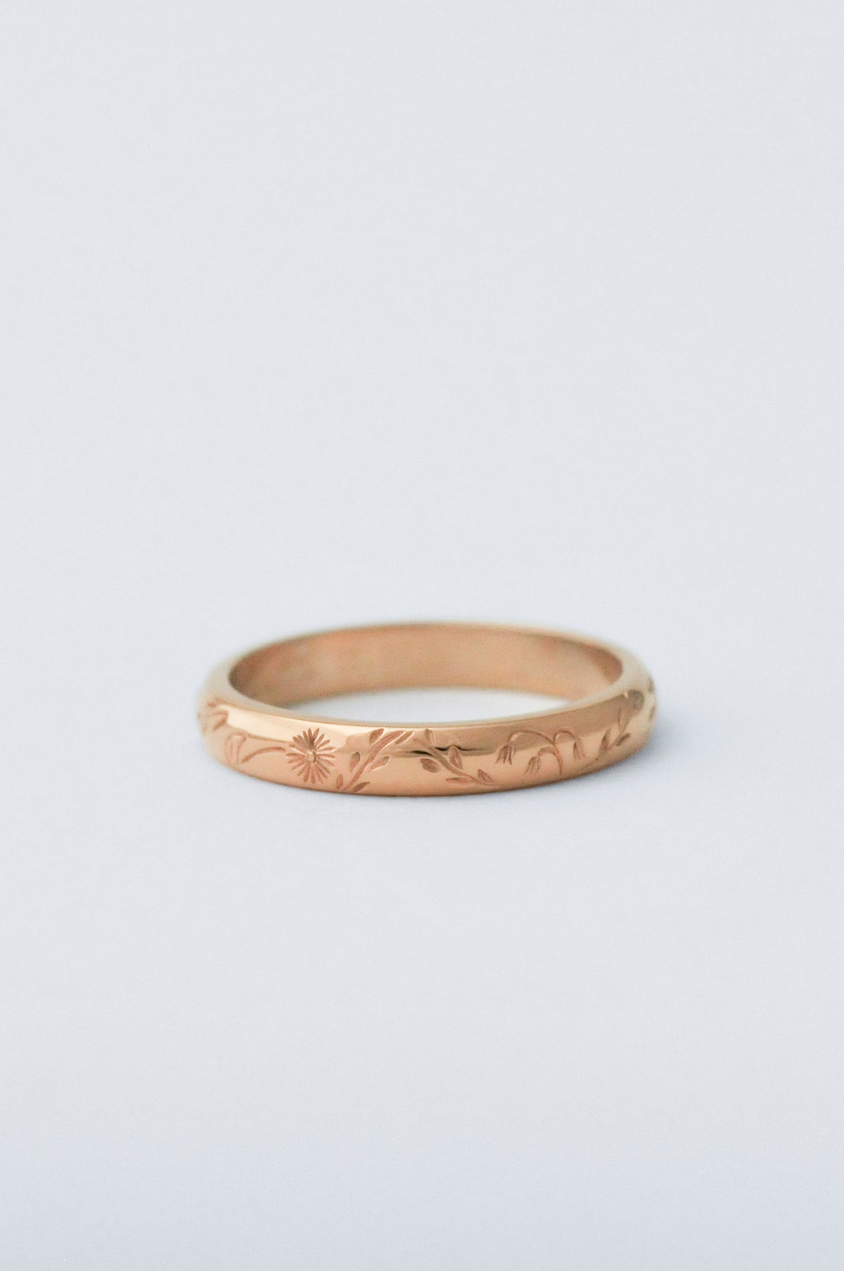 A thin gold band ring with delicate hand engraved florals, photographed on a clean, light gray background.
