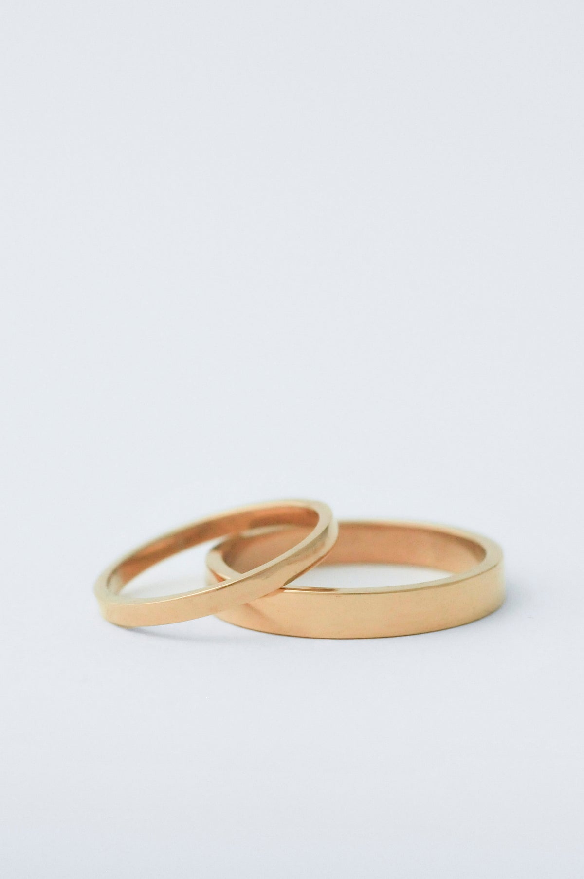 Two minimalist gold wedding bands, both with a flat face, one s 2mm the other 5mm resting on a clean white surface.