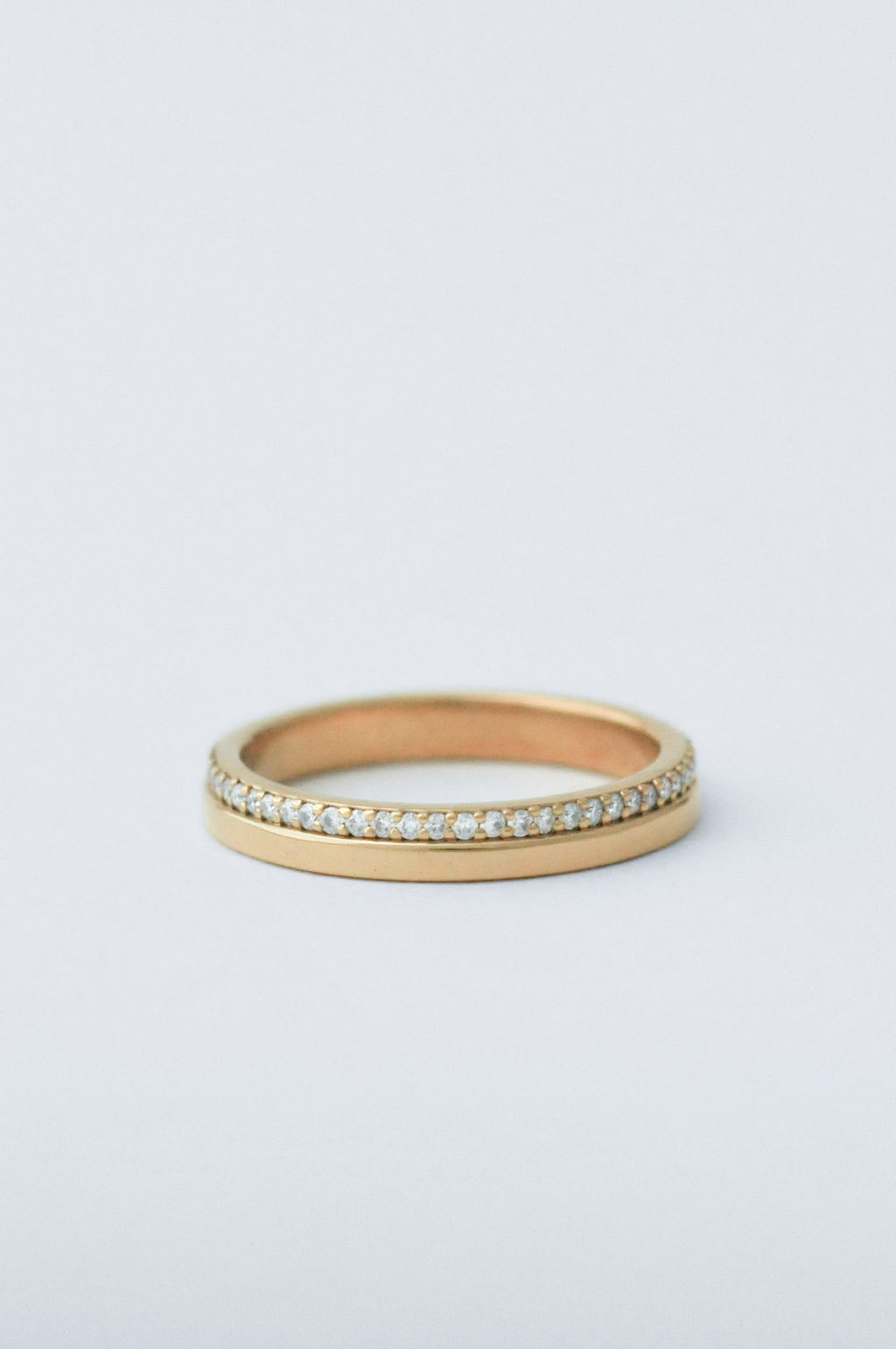 A thin yellow gold eternity ring featuring a channel-set row of small clear stones on the outer band, photographed against a seamless light gray background.