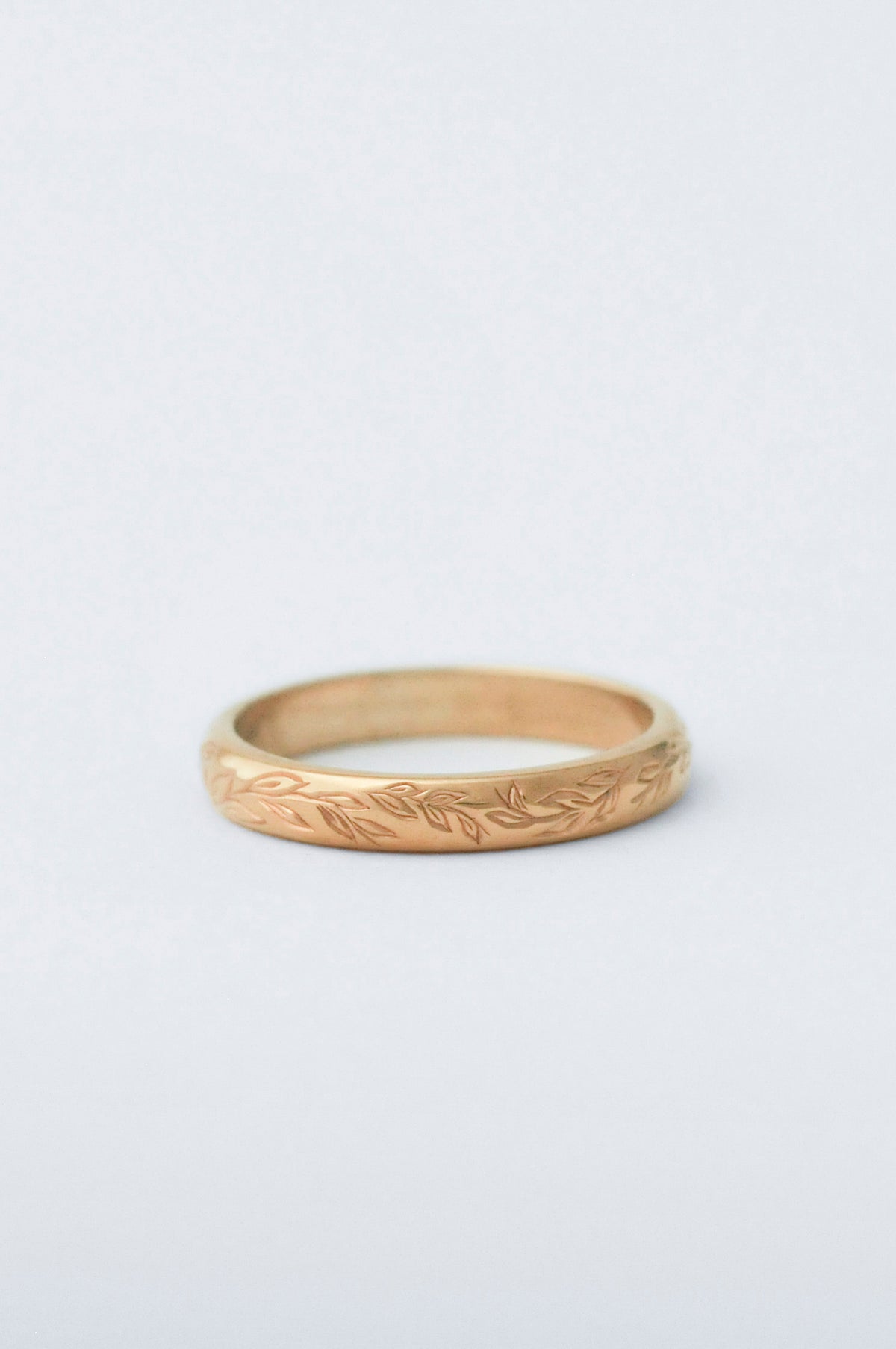 Gold wedding band ring with willow leaf hand engraved detailing on a light grey background
