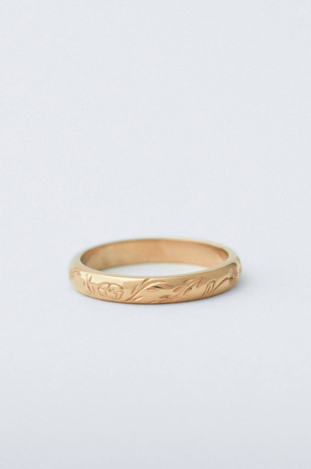 A yellow gold band ring with a delicate, low-profile floral engraving, photographed on a clean, light gray background.