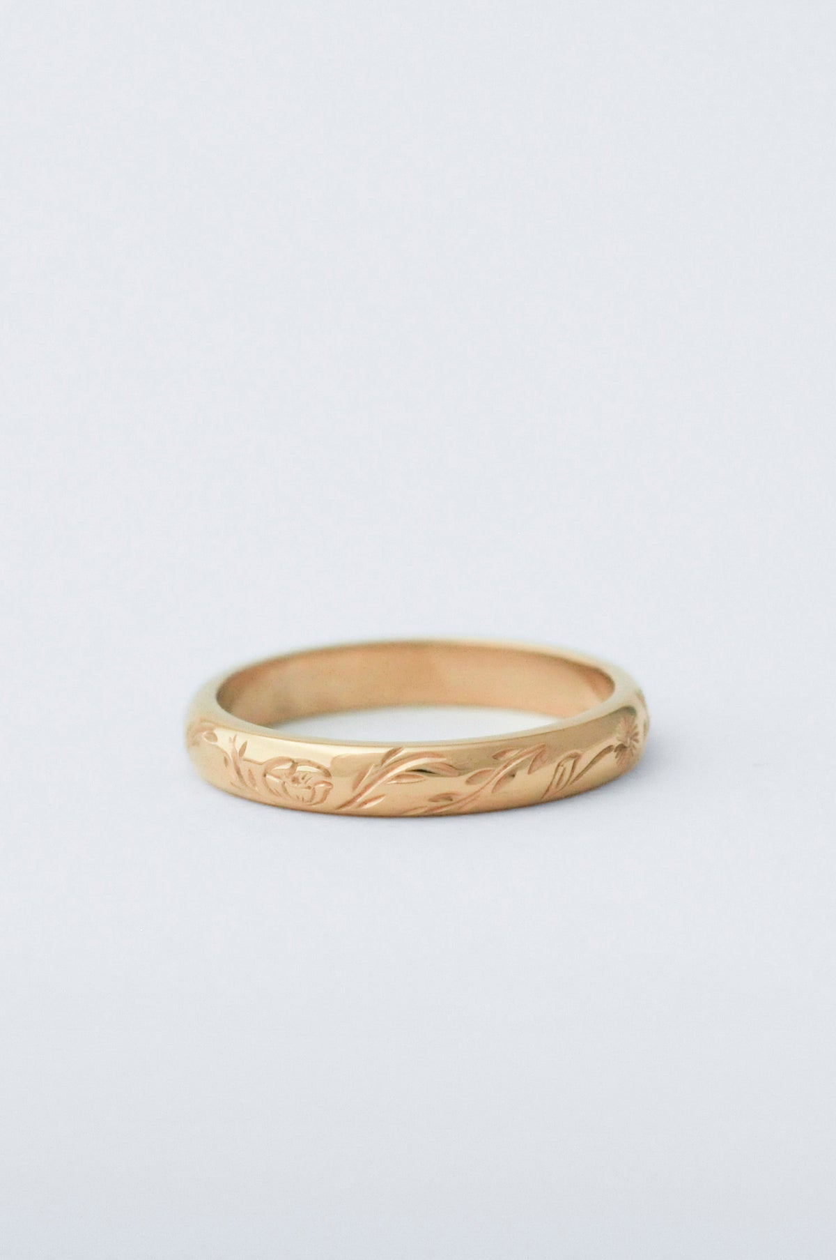A yellow gold band ring with a delicate, low-profile floral engraving, photographed on a clean, light gray background.