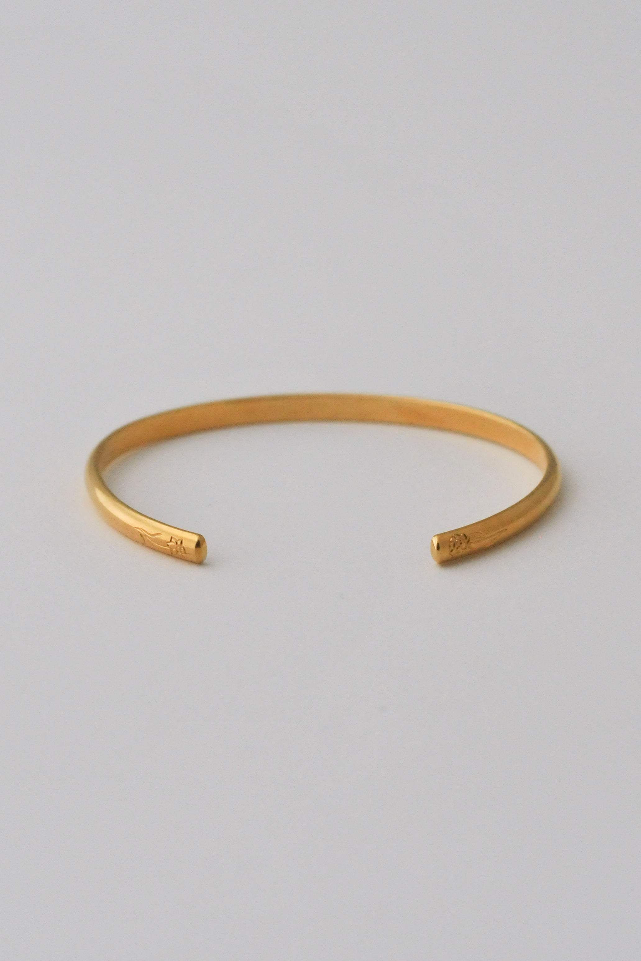 A minimalist vermeil gold open cuff bangle bracelet with hand engraved flowers on each end, photographed on a clean, white background.