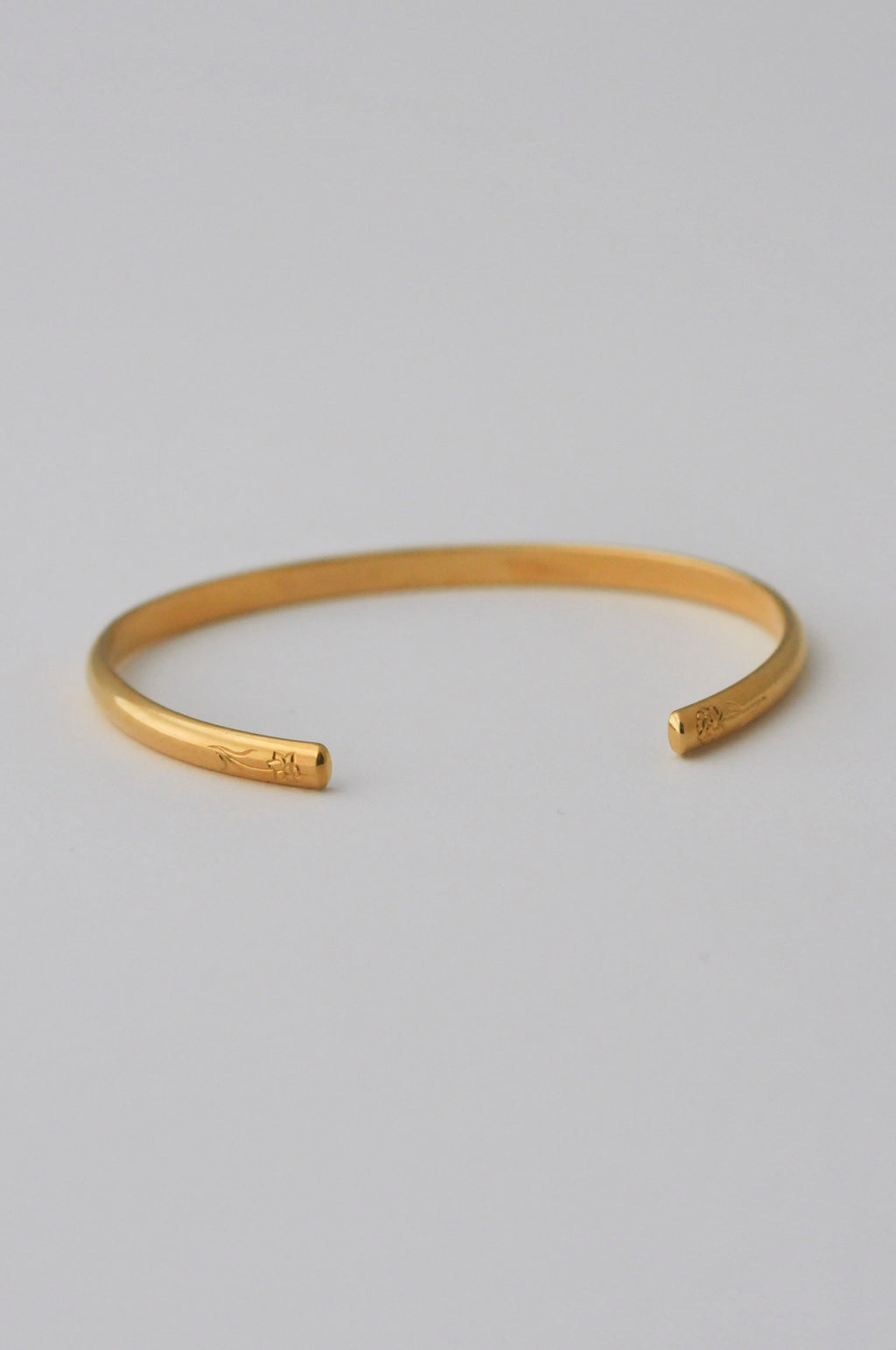A minimalist vermeil gold open cuff bangle bracelet with hand engraved flowers on each end, photographed on a clean, white background.