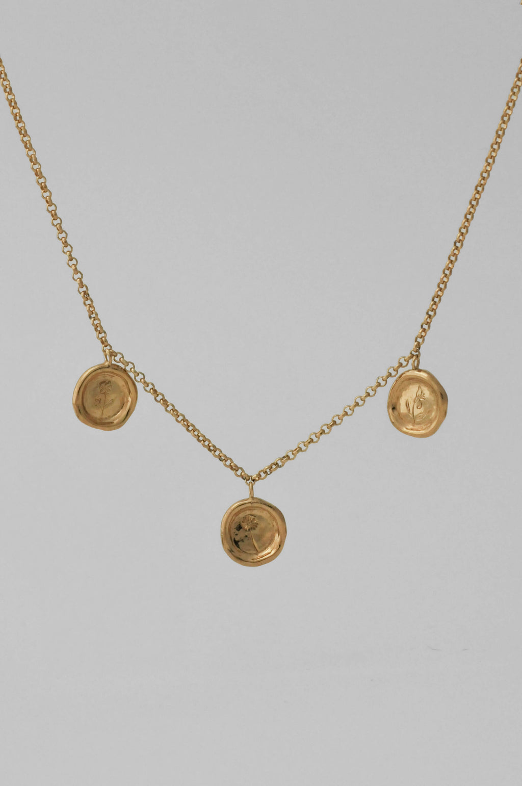 A gold necklace with three circular wax-sealed styled pendant charms with single hand engraved flowers, displayed against a solid light gray background.