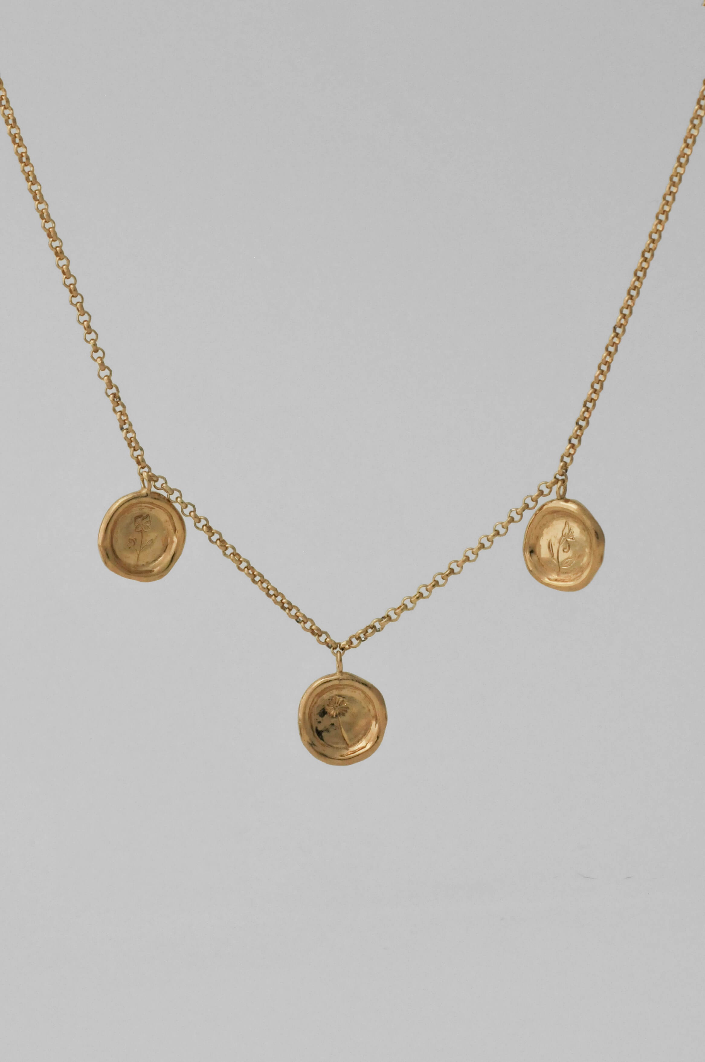 A gold necklace with three circular wax-sealed styled pendant charms with single hand engraved flowers, displayed against a solid light gray background.
