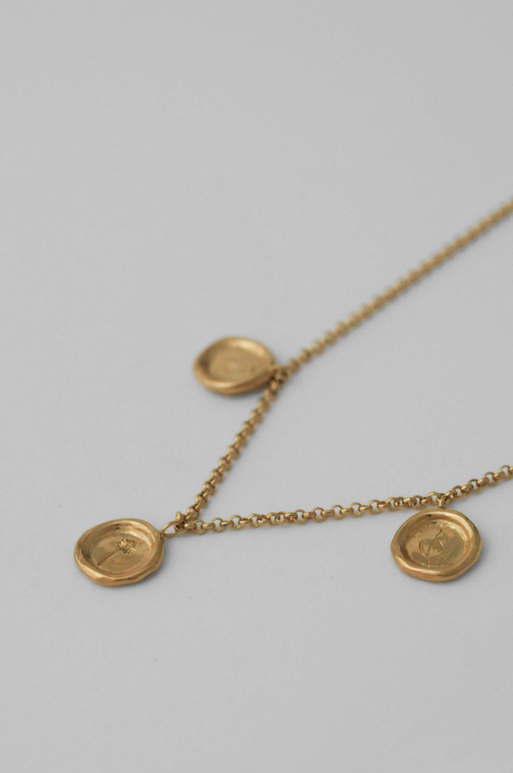 A delicate gold chain necklace featuring three small, round, wax-sealed styled pendants with hand engraved flowers, displayed on a neutral light gray background.