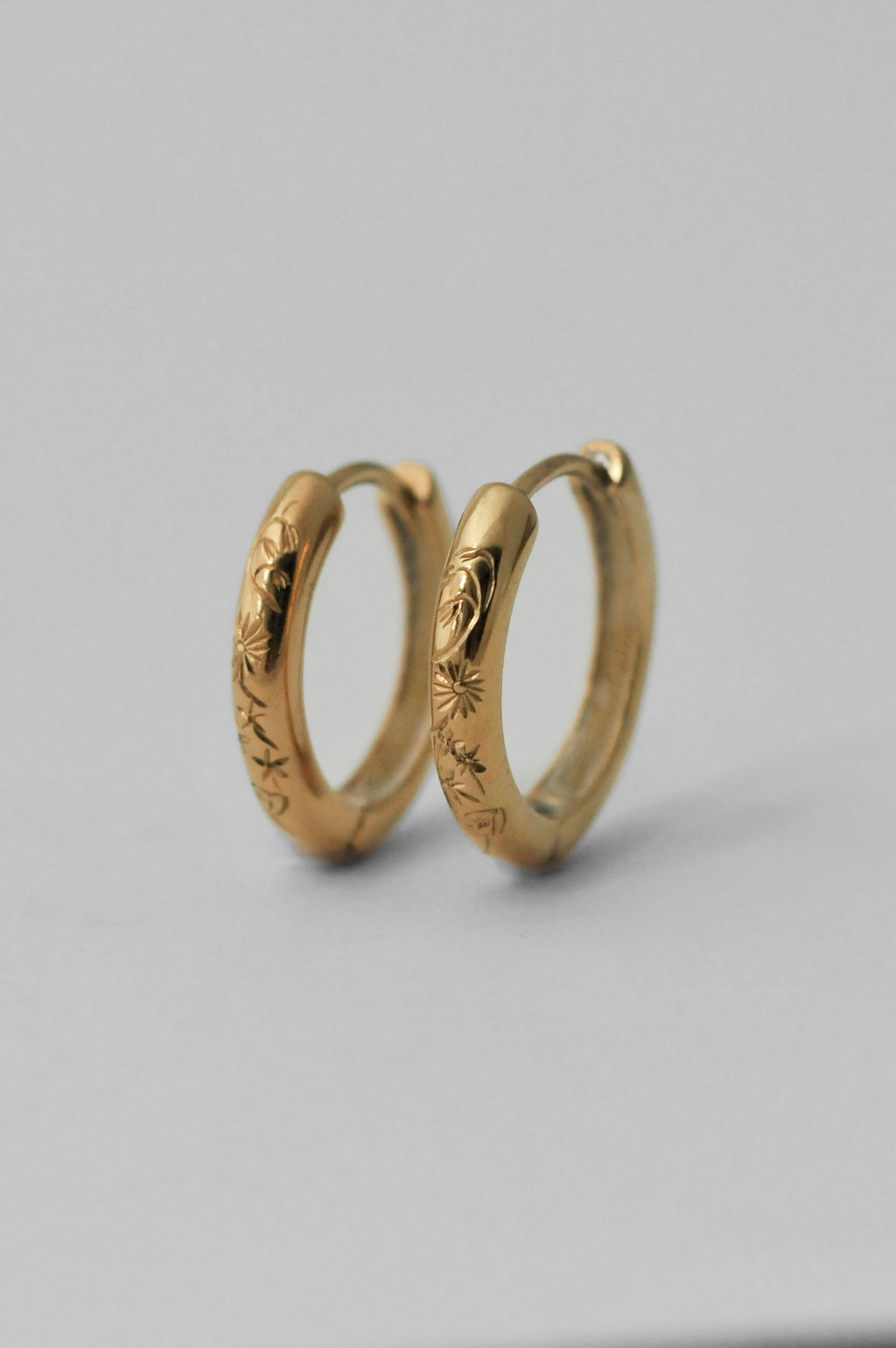 A pair of small gold huggie hoop earrings with hand engraved floral patterns, displayed on a neutral light gray background.