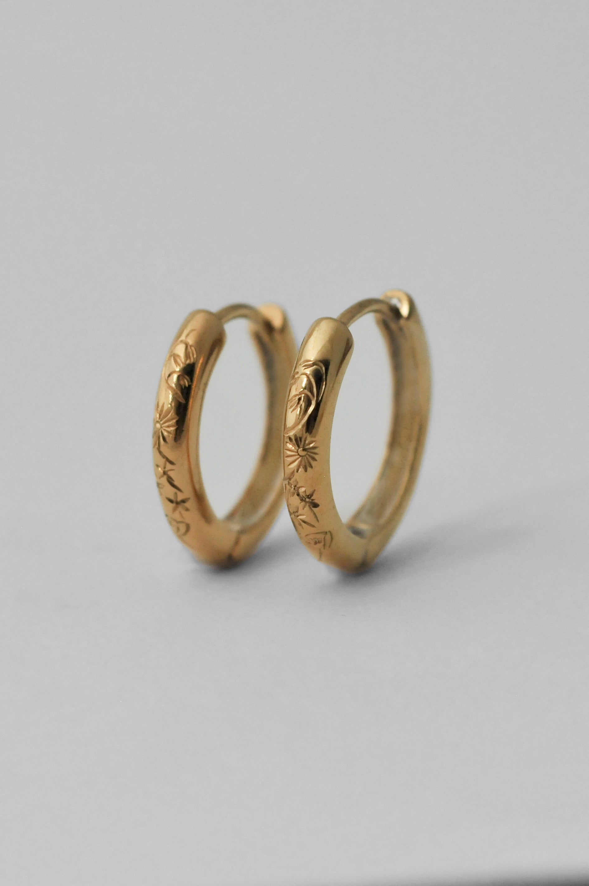 A pair of small gold huggie hoop earrings with hand engraved floral patterns, displayed on a neutral light gray background.