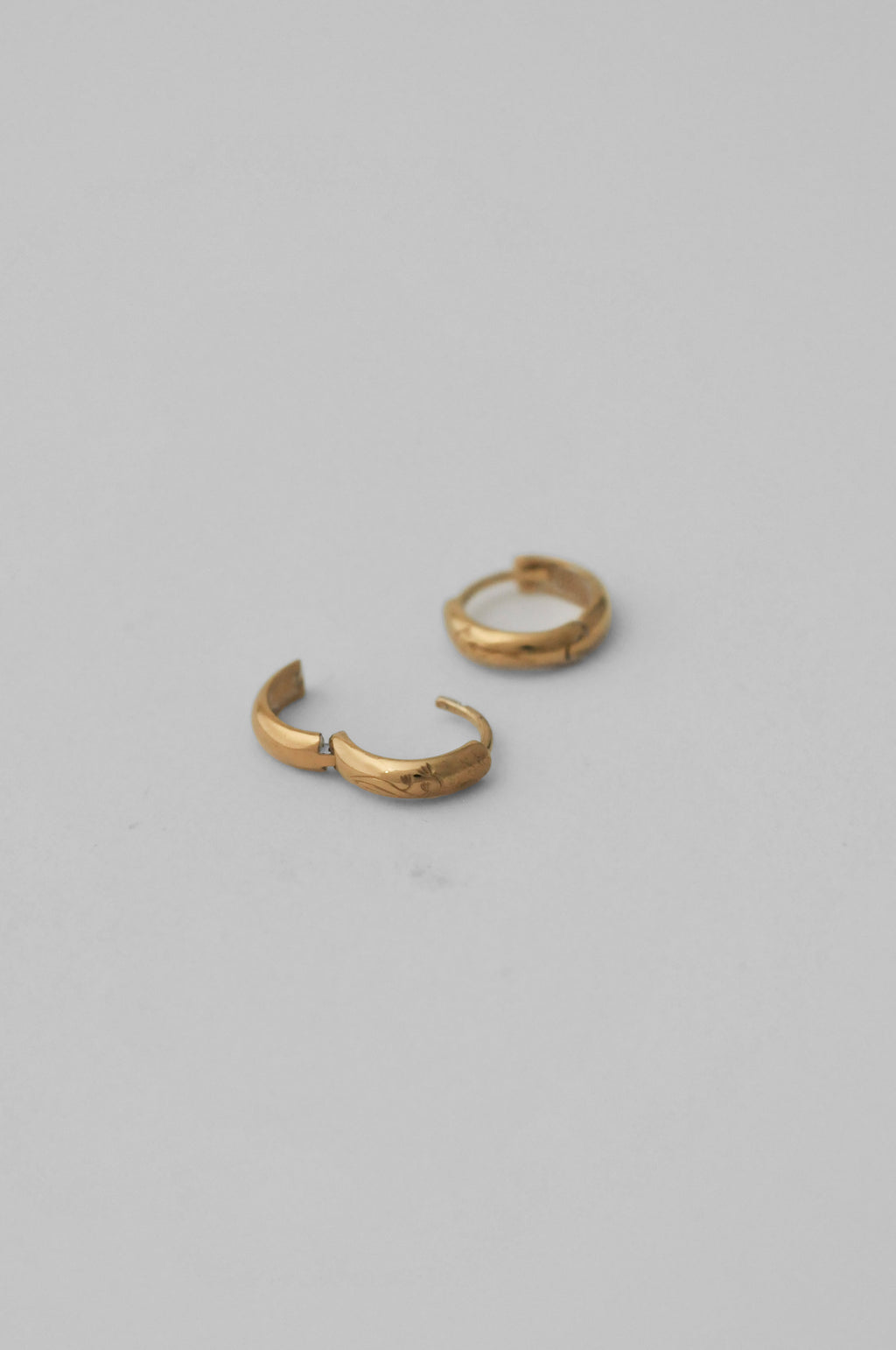 A pair of small gold huggie hoop earrings with hand engraved floral details, showing the opening of the earring, shown on a neutral gray background