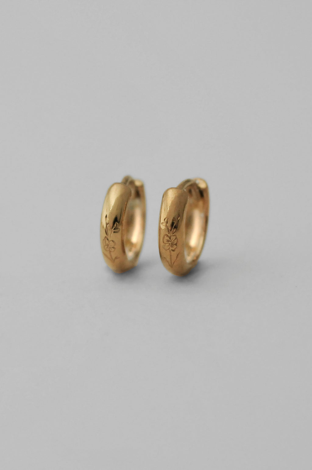 A pair of small gold huggie hoop earrings with hand engraved floral details, shown on a neutral gray background.