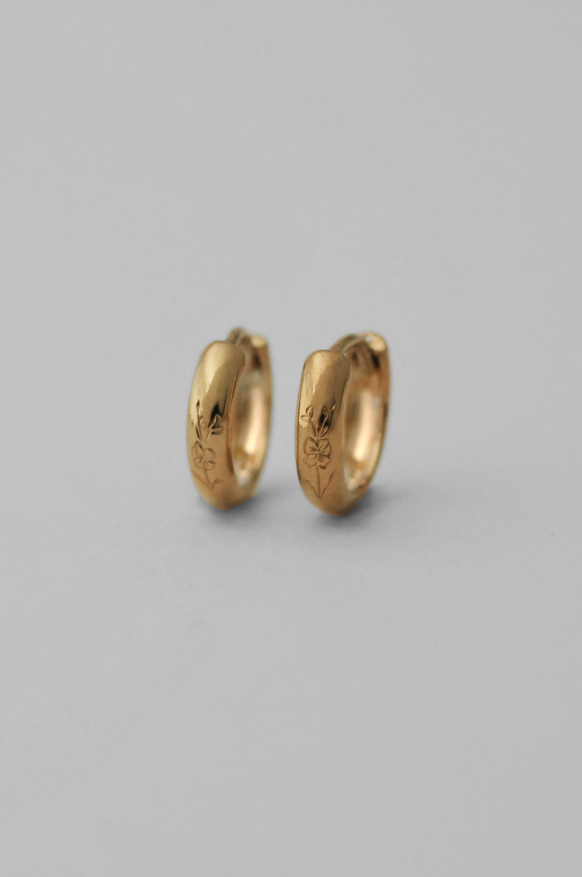 A pair of small gold huggie hoop earrings with hand engraved floral details, shown on a neutral gray background.