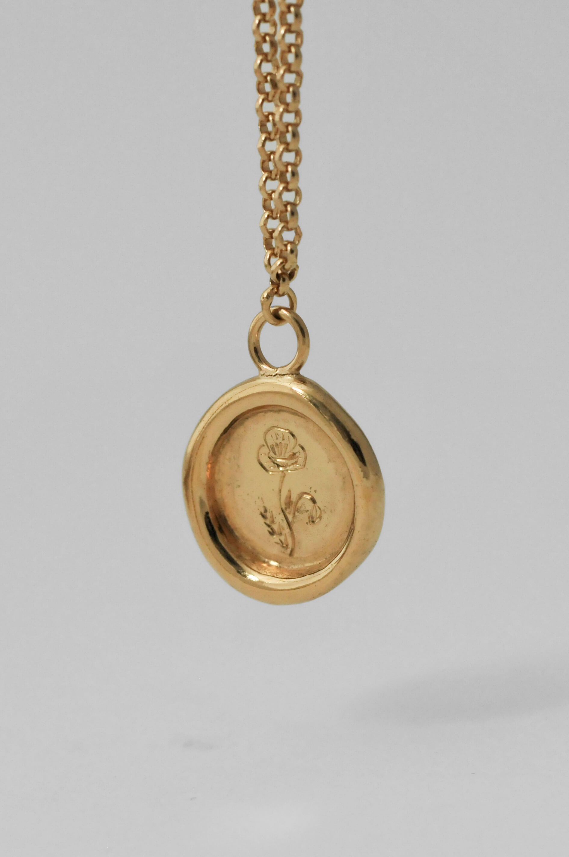 A gold pendant necklace featuring a circular wax-seal style charm with an engraved birth flower design, suspended from a gold chain against a neutral light gray background.