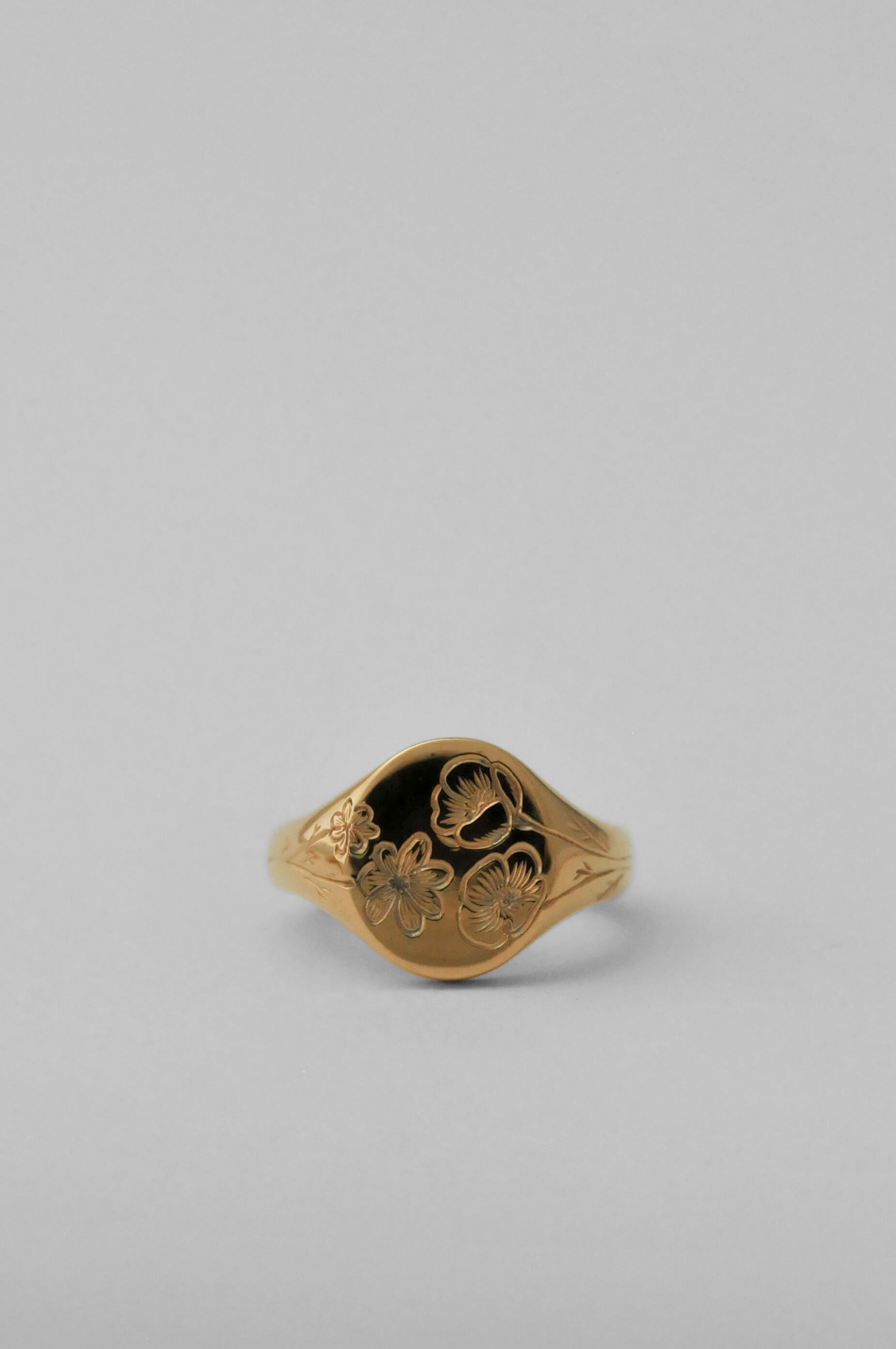 A shiny gold oval signet ring with an engraved birth flower floral pattern, set against a solid light grey background.