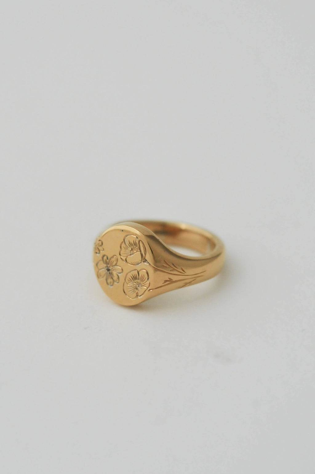 A gold oval pinky signet ring with bespoke birth flower engraving set against a solid light grey background.