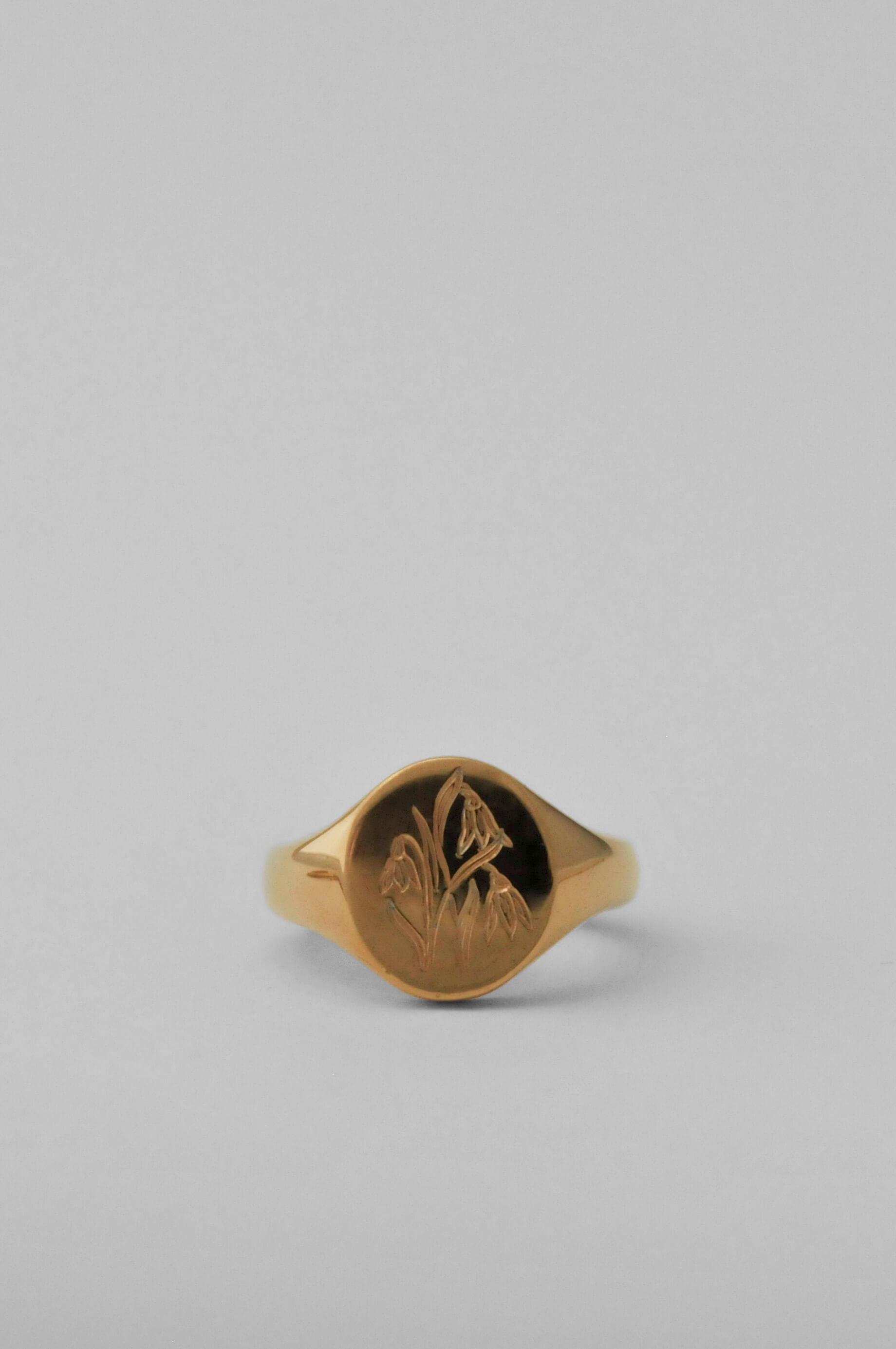 A gold signet ring with an engraved floral motif on a smooth, polished band, set against a solid light grey background.