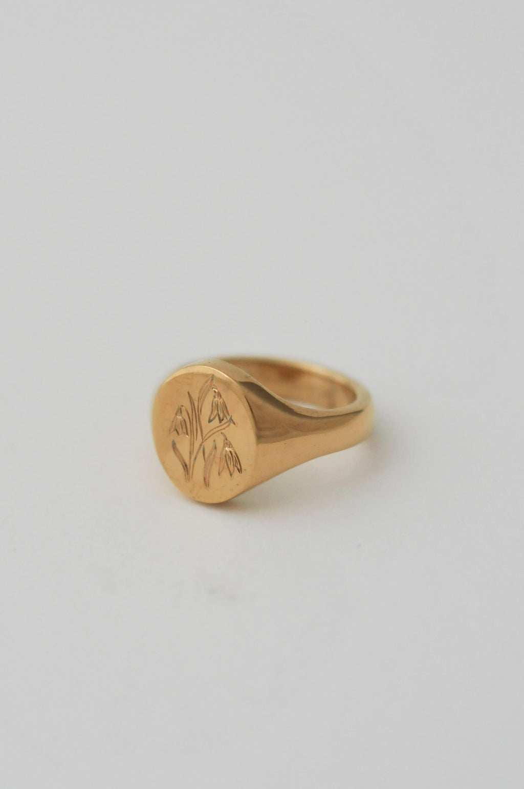A gold signet ring with an engraved floral motif on a smooth, personalised birth flower engraving, set against a solid light grey background.