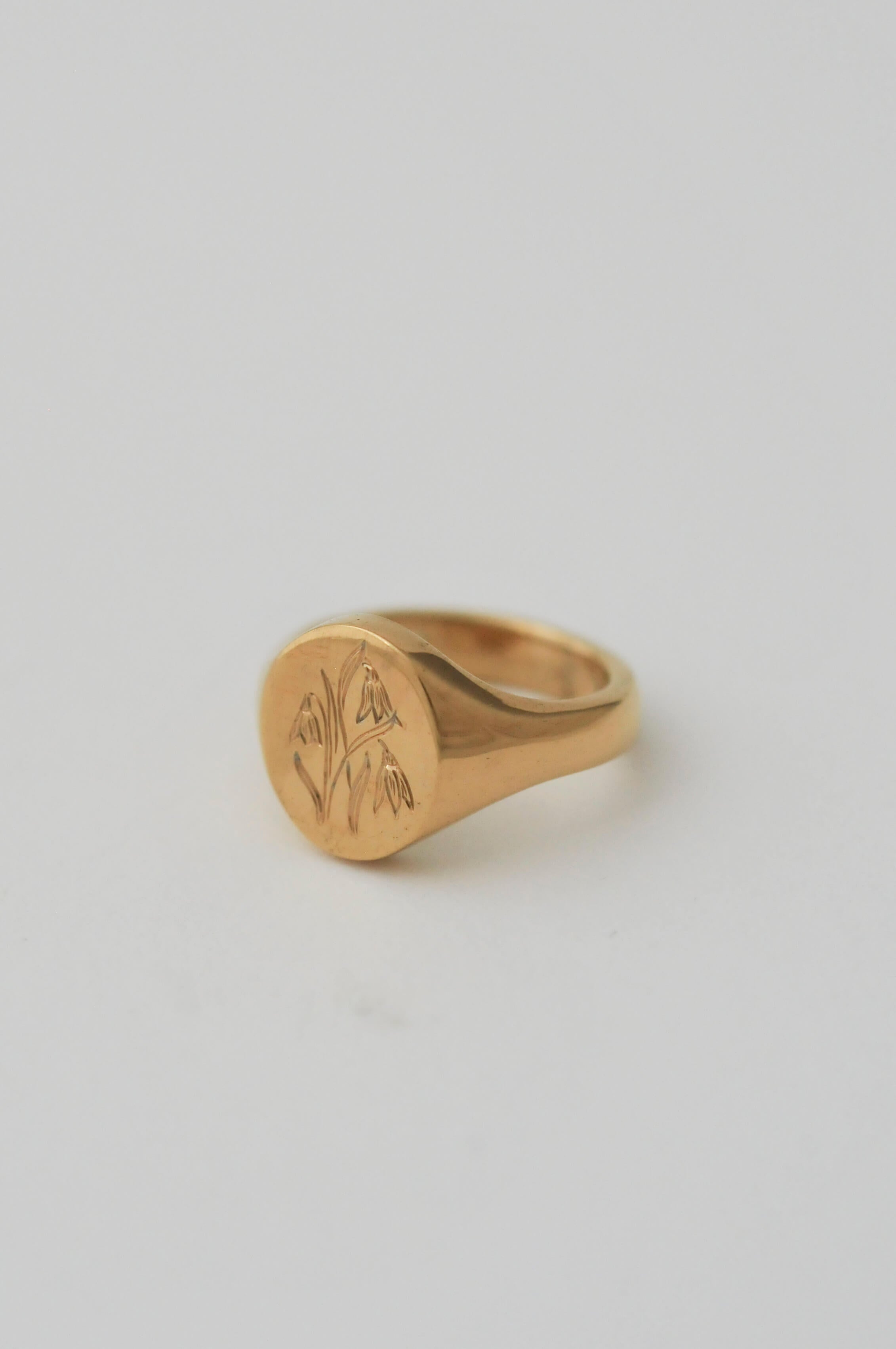 A gold signet ring with an engraved floral motif on a smooth, personalised birth flower engraving, set against a solid light grey background.