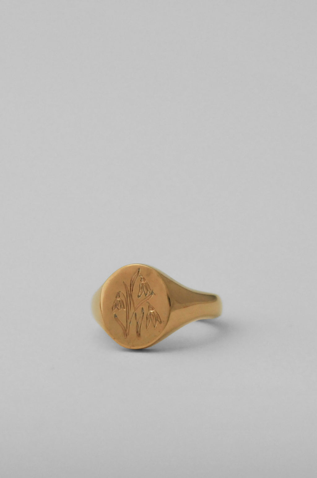A gold signet ring with an engraved floral motif on a smooth, personalised birth flower engraving, set against a solid light grey background.
