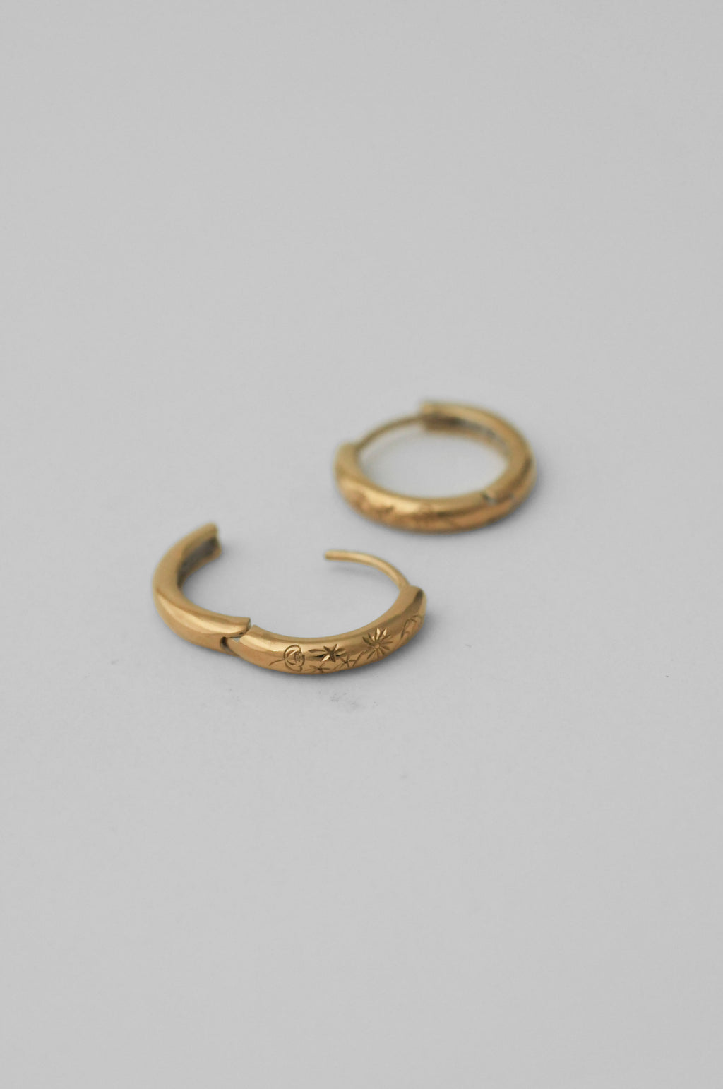A pair of small hand engraved floral gold huggie hoop earrings on a plain light gray background.